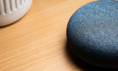 Persona sostiene un smartphone con la app Google Home frente a una lavadora de carga frontal y un altavoz Google Home en la repisa, ilustrando Conectar lavadora a Google Home: guía completa para controlarla por voz.