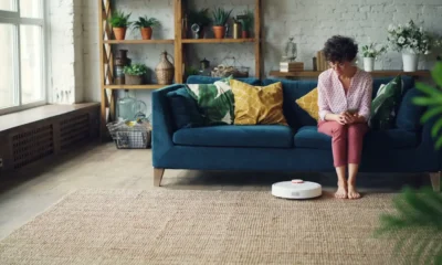 Robot aspirador Dreame Mova E20: análisis, precio y claves — robot aspirador moderno en un salón junto a un sofá sobre suelo de madera, con un teléfono en primer plano mostrando la app de control.