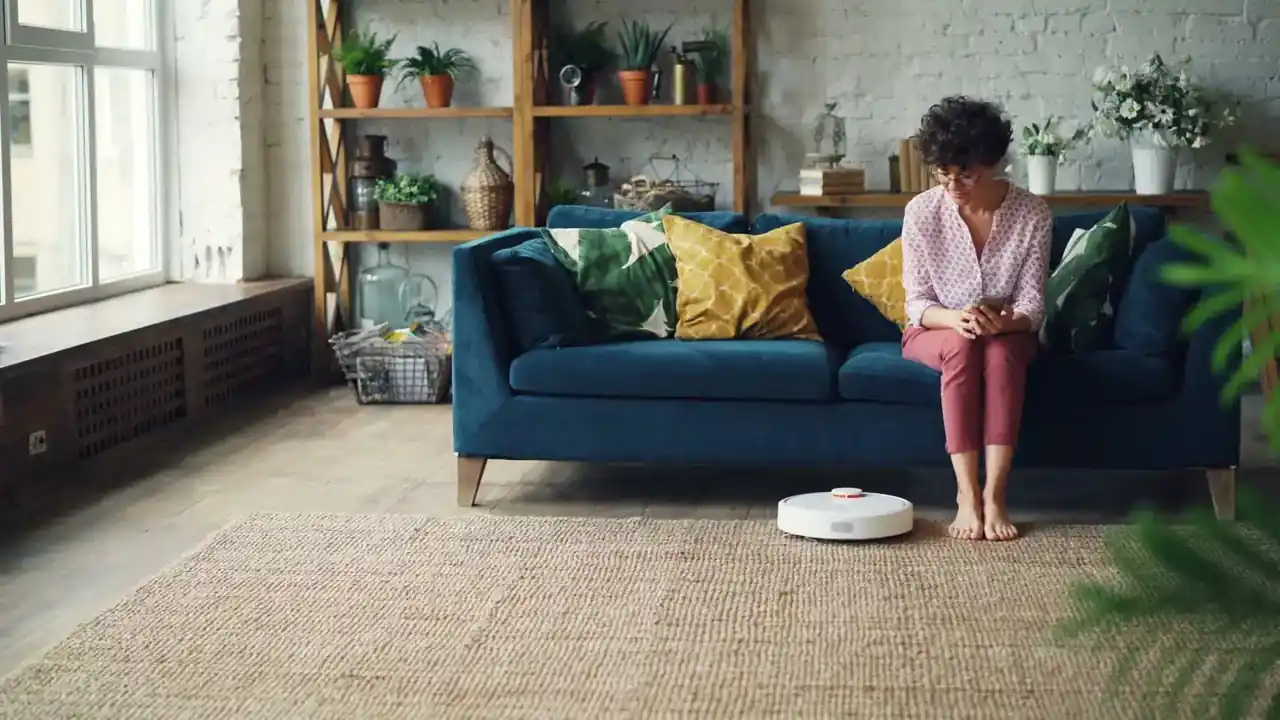 Robot aspirador Dreame Mova E20: análisis, precio y claves — robot aspirador moderno en un salón junto a un sofá sobre suelo de madera, con un teléfono en primer plano mostrando la app de control.