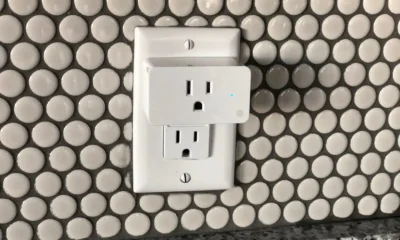 Medidor de consumo enchufable con Wi-Fi: cómo elegirlo bien — primer plano de un medidor enchufable conectado a un tomacorriente, una lámpara encendida y un smartphone mostrando la app con consumo eléctrico en tiempo real.
