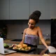 Mujer en una cocina moderna usando un horno inteligente con pantalla táctil que muestra iconos de comida y un plato asado en su interior, ilustrando Hornos inteligentes que identifican la comida y cocinan solos.